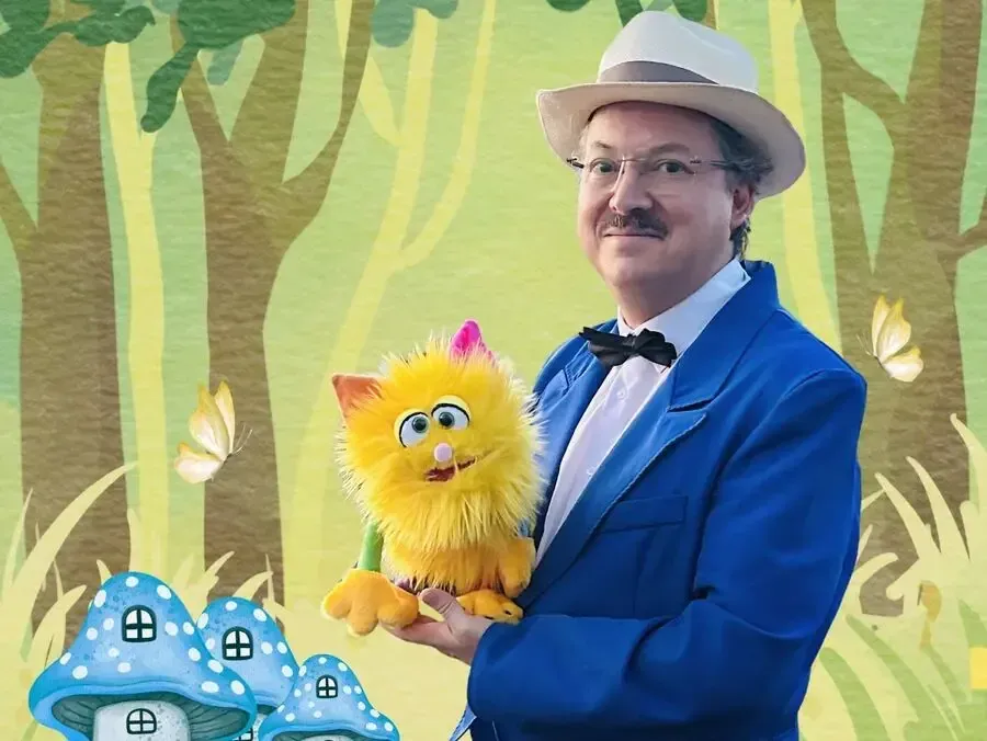 Sylvio, en veste bleue et nœud papillon, tient une marionnette jaune devant un décor de forêt illustré avec des champignons