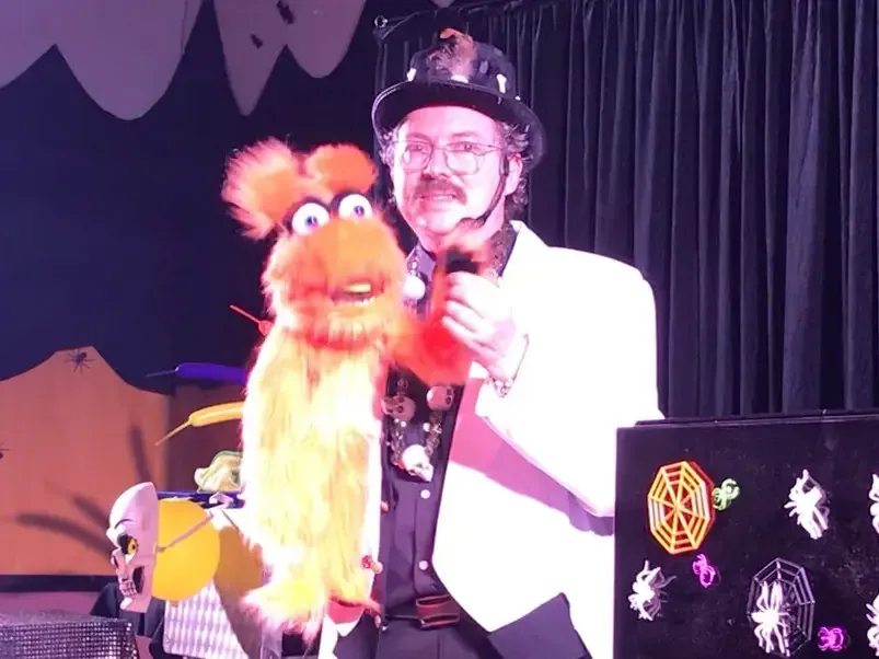 Sylvio en veste blanche et chapeau noir, une marionnette orange à la main, dans le décor du spectacle d’Halloween