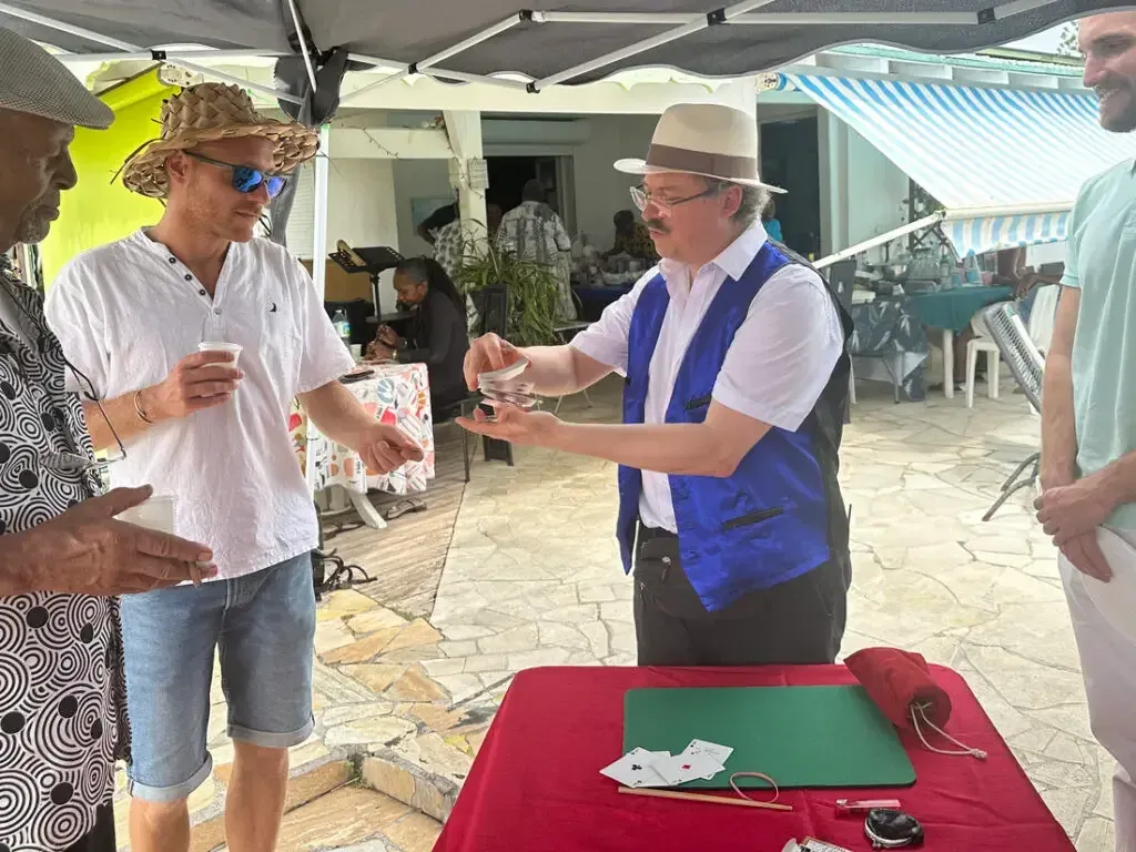 Sylvio, en gilet bleu et chapeau panama, présente un tour à deux invités sous un auvent, sur une table à nappe rouge