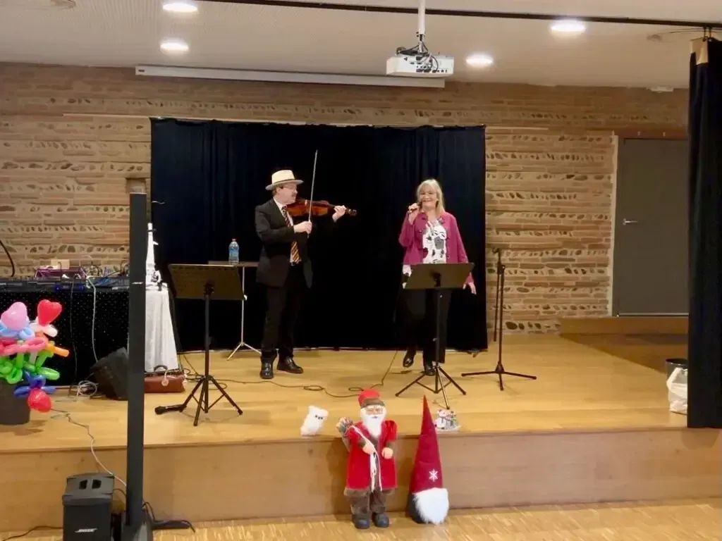 Sylvio au violon et Nathalie au chant sur une scène en bois devant un mur de briques, avec deux figurines de Père Noël au premier plan