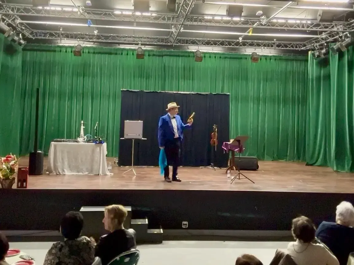 Sylvio, en queue de pie bleue et chapeau panama, sur une scène aux rideaux verts, une baguette à la main