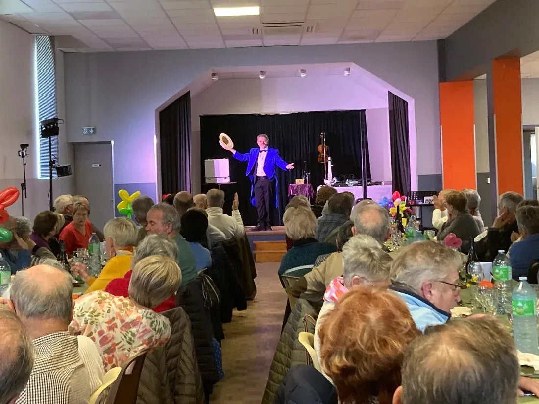 Sylvio, bras ouverts et chapeau levé, sur la scène d’une salle polyvalente, devant des convives attablés vus de dos