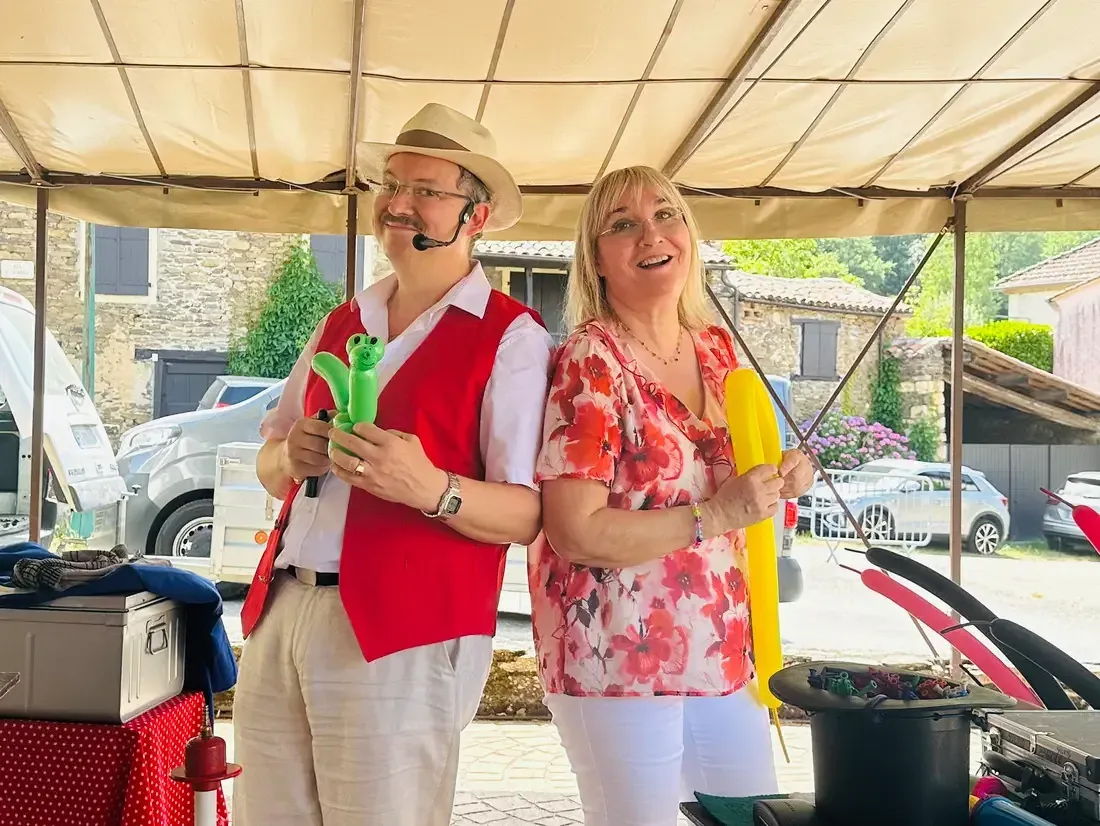 Sylvio, en gilet rouge, et Nathalie présentent des sculptures sur ballons sous une tonnelle